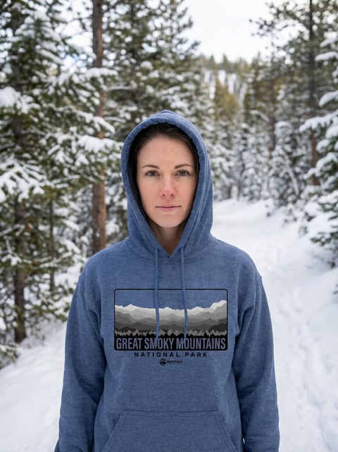 Woman wearing blue Great Smoky Mountains hoodie standing in snowy forest with evergreen trees