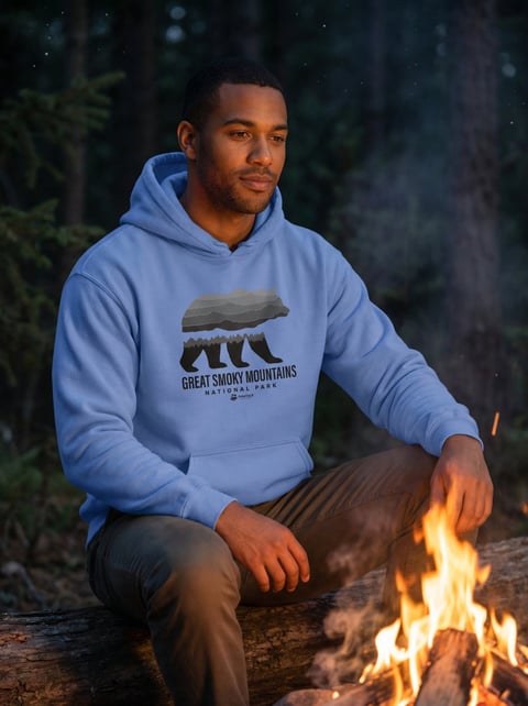 Man in blue Great Smoky Mountains hoodie crouching by campfire at night in forest