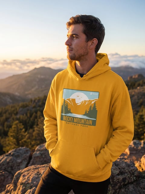 Man wearing yellow Rocky Mountain hoodie standing on mountain overlook at sunset with forested mountains in background